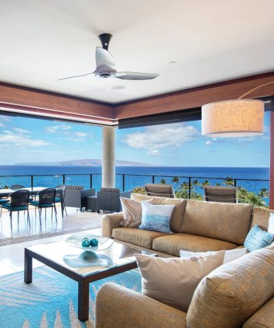 Stylish coastal living room opens to a sea-view balcony with a beige sofa, glass coffee table, wicker chairs, and a ceiling fan, bright and airy.