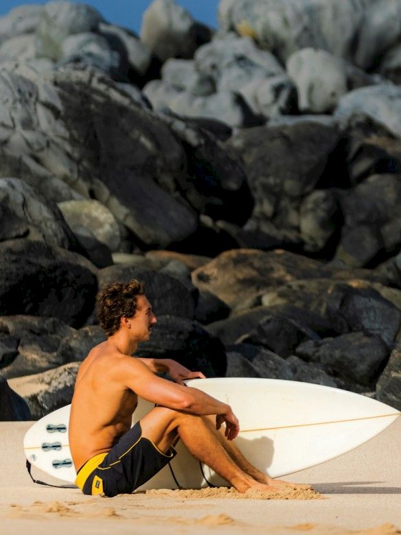 A shirtless man sits on a sandy beach with a surfboard, rocks in the background, calm vibe, prepping or resting after surfing.