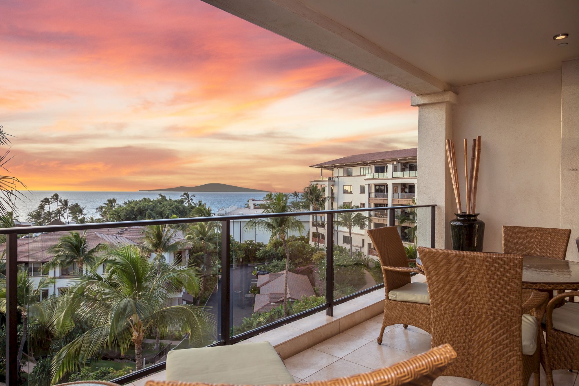 A balcony with wicker furniture overlooks palm trees and a sunset over the ocean, with a neighboring building in the distance, creating a tropical view.