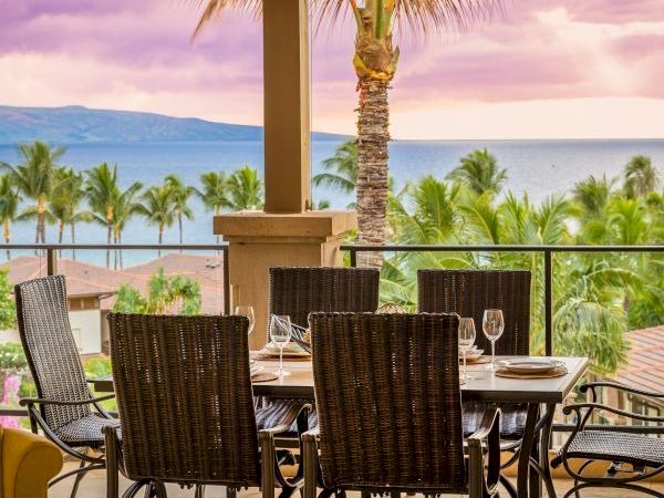 A beachfront patio with a dining table set for eight, wicker chairs, palm trees, sunny sky, and an ocean view in the background.