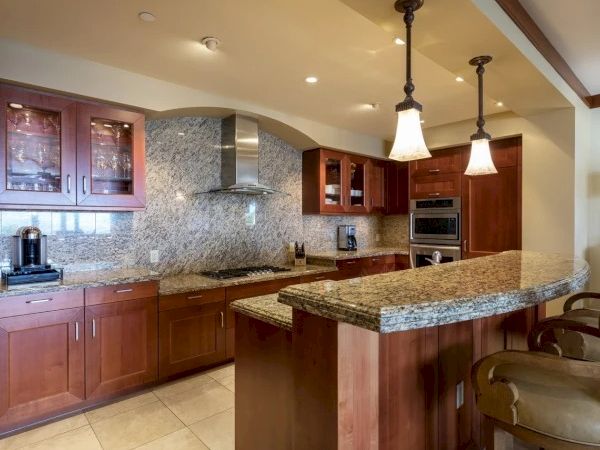 A modern kitchen with granite countertops, warm wood cabinets, a tiled backsplash, stainless appliances, and a curved breakfast bar with hanging lights.