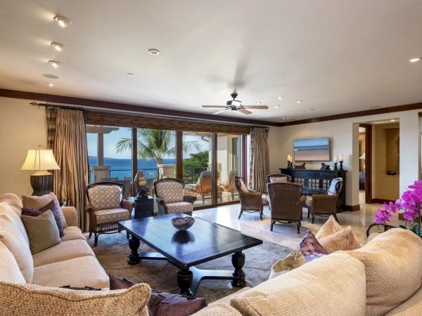 A bright, cozy living room with beige sofas, a dark wood coffee table, a ceiling fan, and floor-to-ceiling windows revealing a sunny view outside, ending with a period.