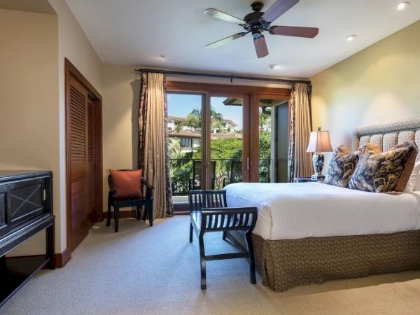 A bright hotel room with a king bed, ceiling fan, balcony doors, two nightstands, a desk, and a chair, looking out onto a patio.