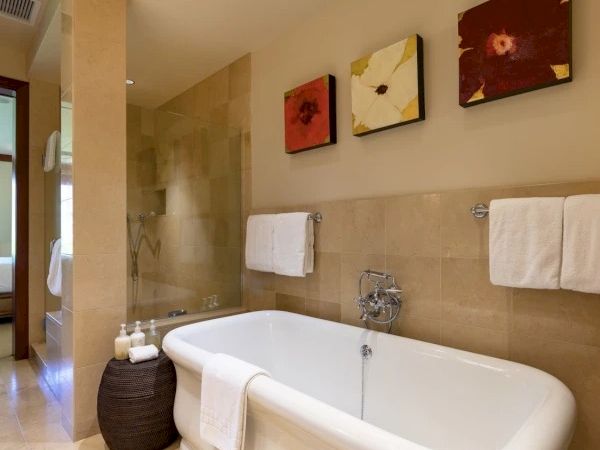 A modern bathroom with a freestanding oval bathtub, beige tiles, white towels, and three wall art pieces above the tub, plus a glass shower in the background.
