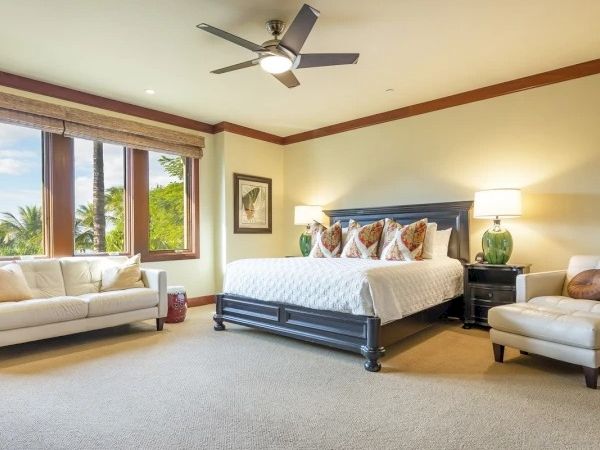 A bright hotel bedroom with a large bed, nightstands, beige carpet, cream sofa and chairs, a ceiling fan, and a big window letting in natural light.