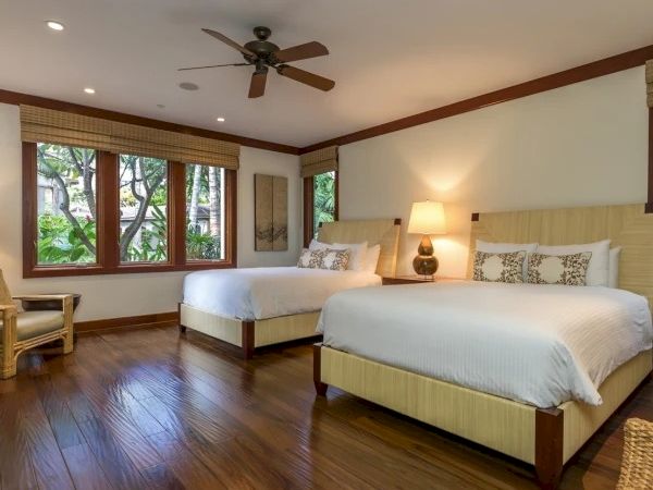 A bright hotel room with two queen beds, wooden floors, a ceiling fan, a large window seat, and a nightstand lamp between the beds.
