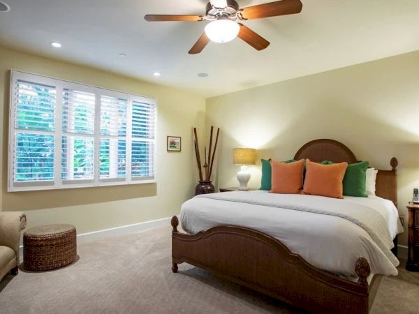 A cozy bedroom with a wooden bed, cream walls, beige carpet, white bedding, colorful throw pillows, a ceiling fan, and large window blinds.
