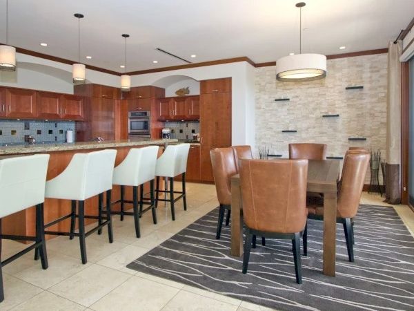 A modern cafe or hotel lounge with a bar area, white and brown seating, high chairs, a kitchen backdrop, and a patterned rug under brown chairs, nice lighting.