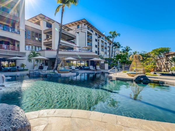 A luxurious hotel pool scene with a clear blue sky, palm trees, sun loungers, and modern white buildings surrounding a large, inviting pool.