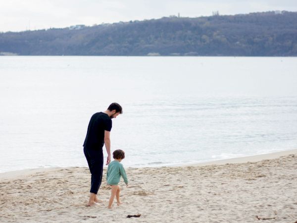 A person and a child walking hand in hand on a sandy beach by the water, with hills in the distance, calm sky, and light breeze.