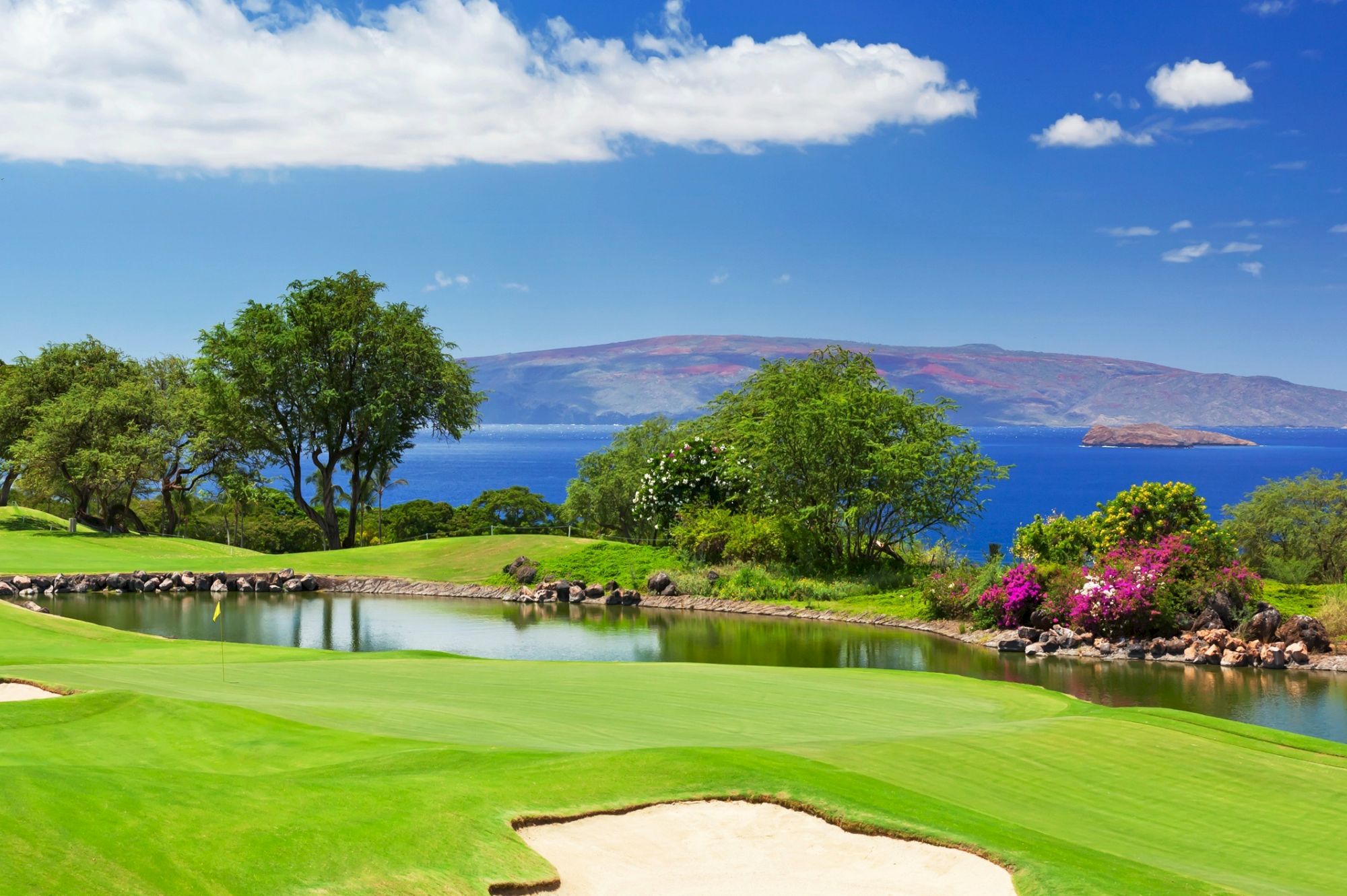 A scenic golf course with lush green fairways, a sand bunker, a calm water hazard, trees, and a backdrop of a sparkling blue lake under a bright sky.