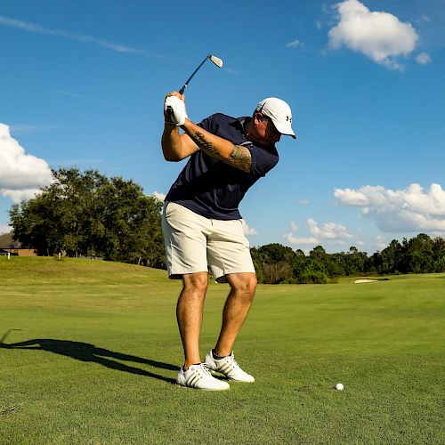 A golfer in a white hat and navy shirt swings a club on a sunny green fairway, with a tee and ball nearby against a blue sky.