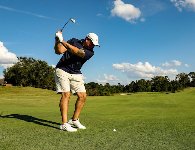 A golfer in a white hat and navy shirt swings a club on a sunny green fairway, with a tee and ball nearby against a blue sky.
