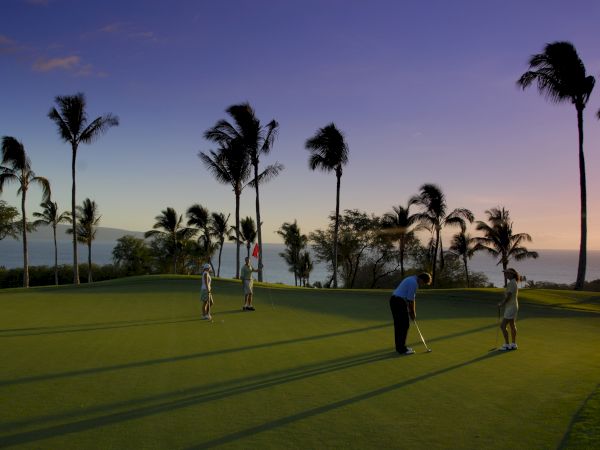A twilight golf scene on a seaside course with palm trees, three players lining up putts, long shadows, and a calm horizon.