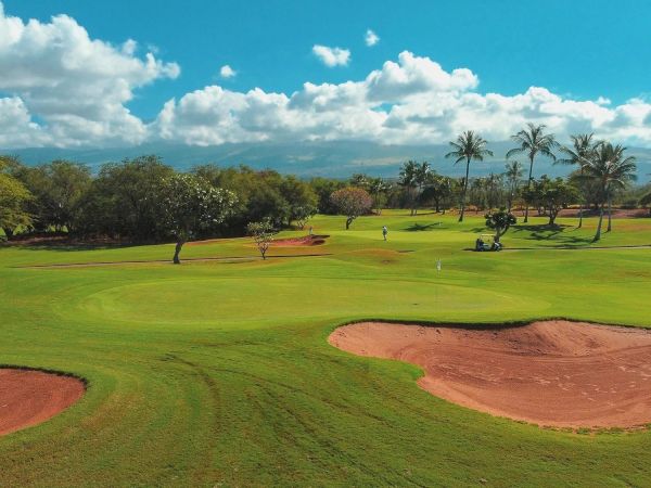 A sunny golf course with green fairways, sand bunkers, and palm trees under a blue, cloudy sky. End.
