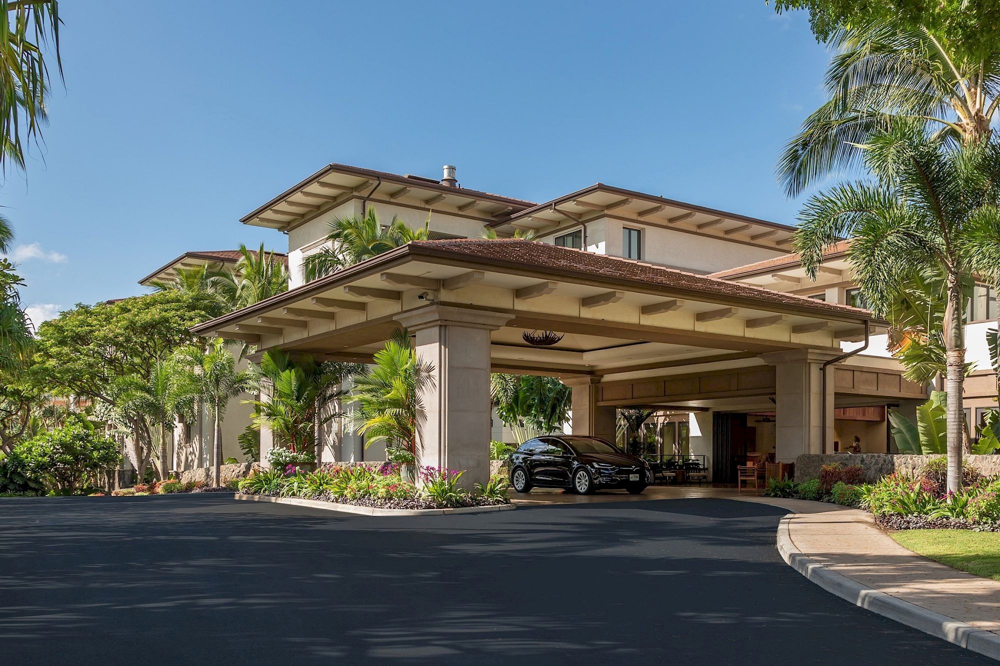 A large hotel or resort entrance with a wide porte-coch&egrave;re, palm trees, and a parked car under shade, sunny day, clean driveway, inviting lobby area.