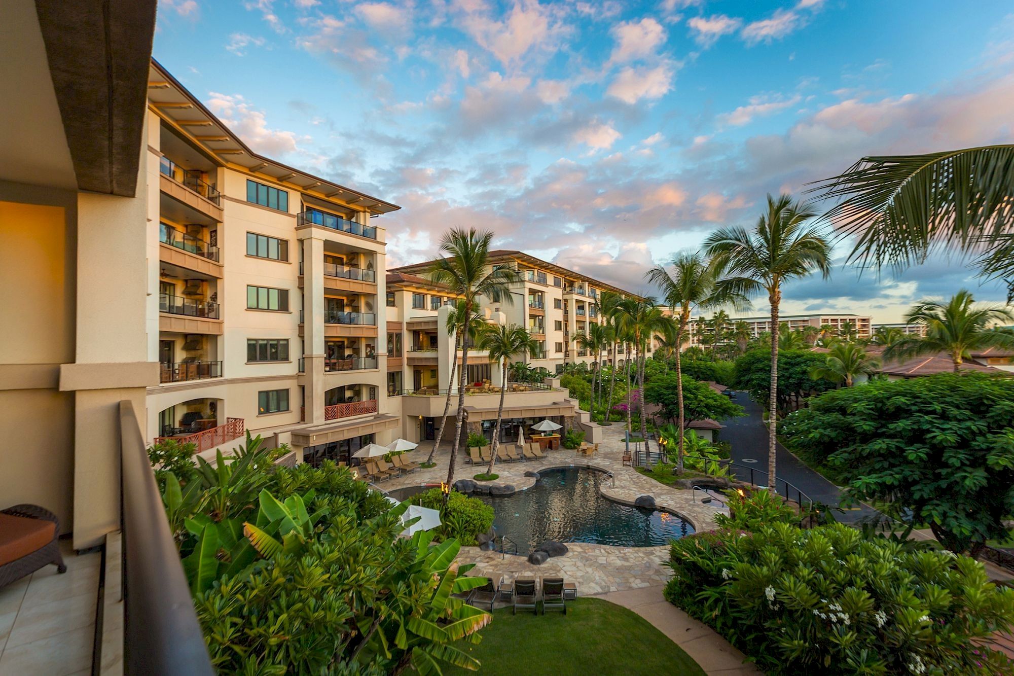 A tropical resort courtyard with palm trees, a pool area, and multi-story hotel buildings under a partly cloudy blue sky.