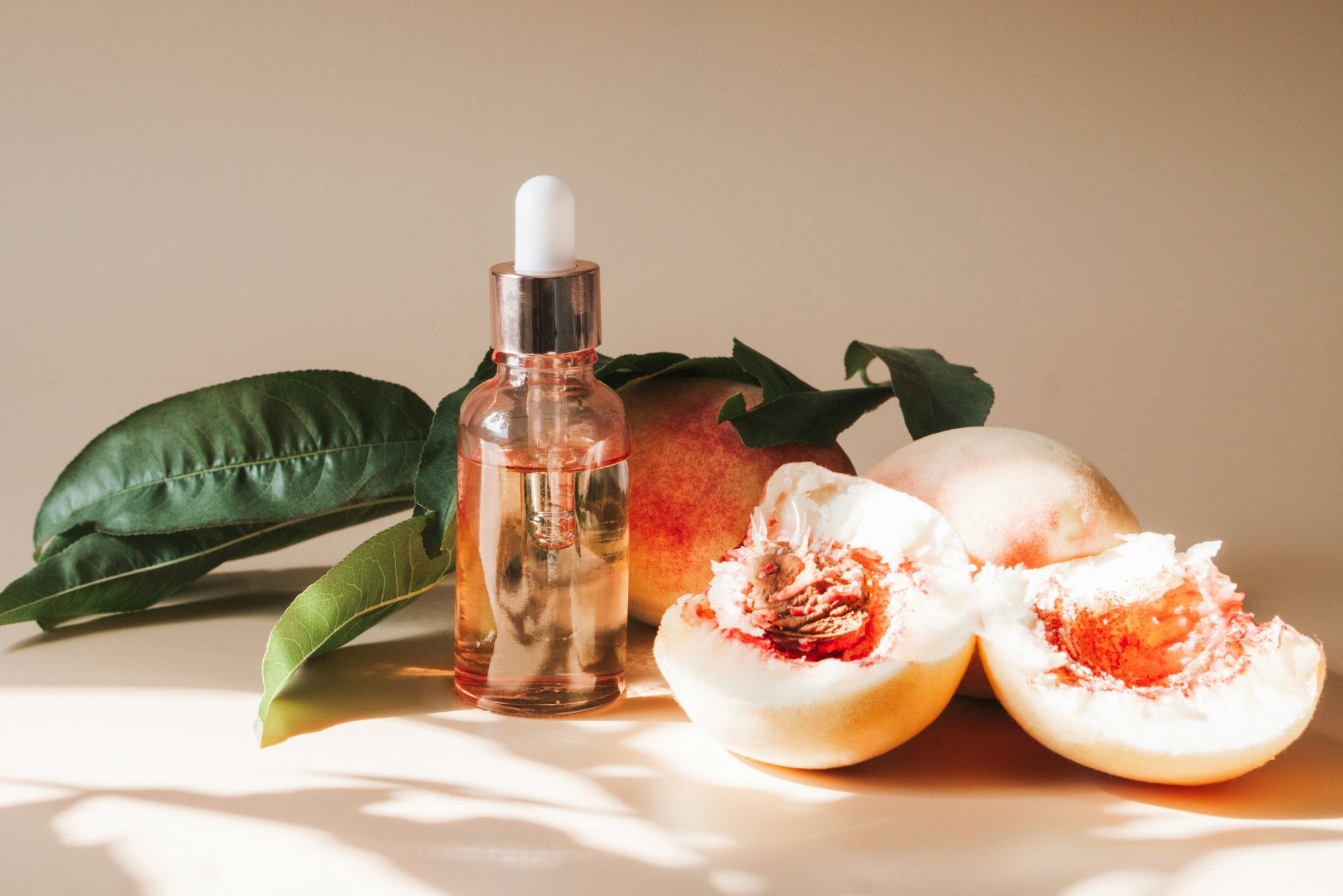 A bottle with a dropper sits beside halved peaches or nectarines and green leaves on a light, sunny surface.
