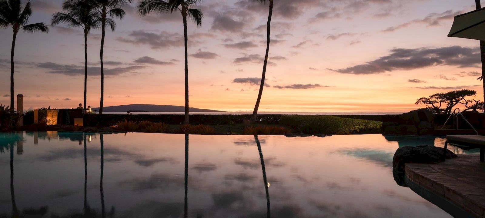 A tranquil tropical sunset by a pool, palm trees silhouette against orange-pink skies, calm water reflecting the scene like glass.