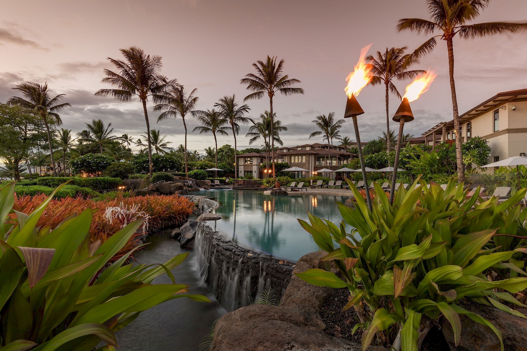 A tropical pool area at sunset with palm trees, lush plants, and tiki torches along a cascading pool, calm water, and resort buildings in the background.