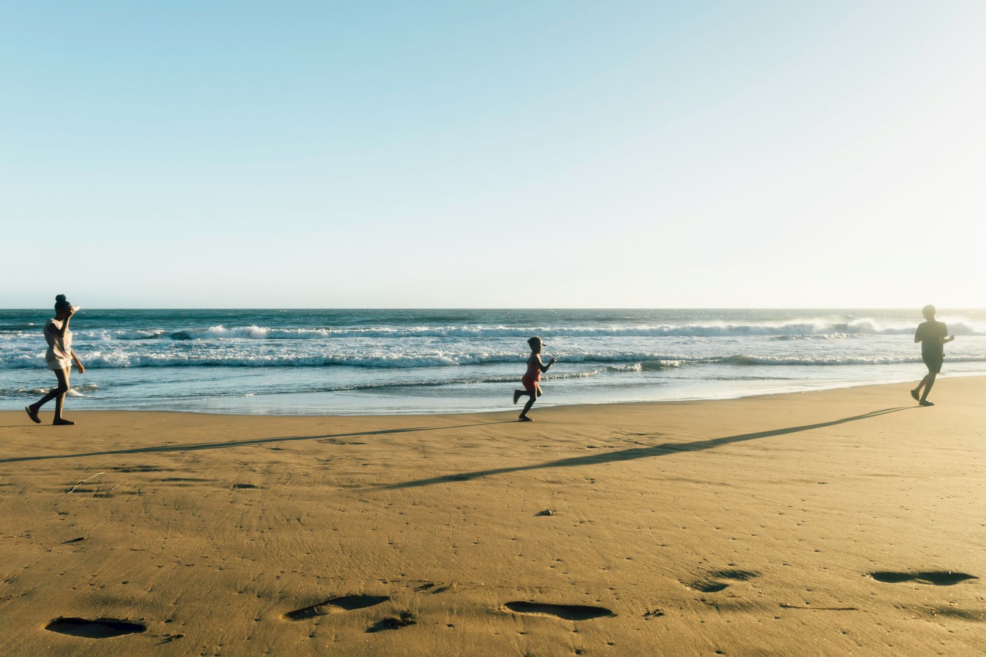 People walking along a sunny beach with the ocean waves, footprints in the sand, and a clear blue sky, capturing a peaceful seaside scene.