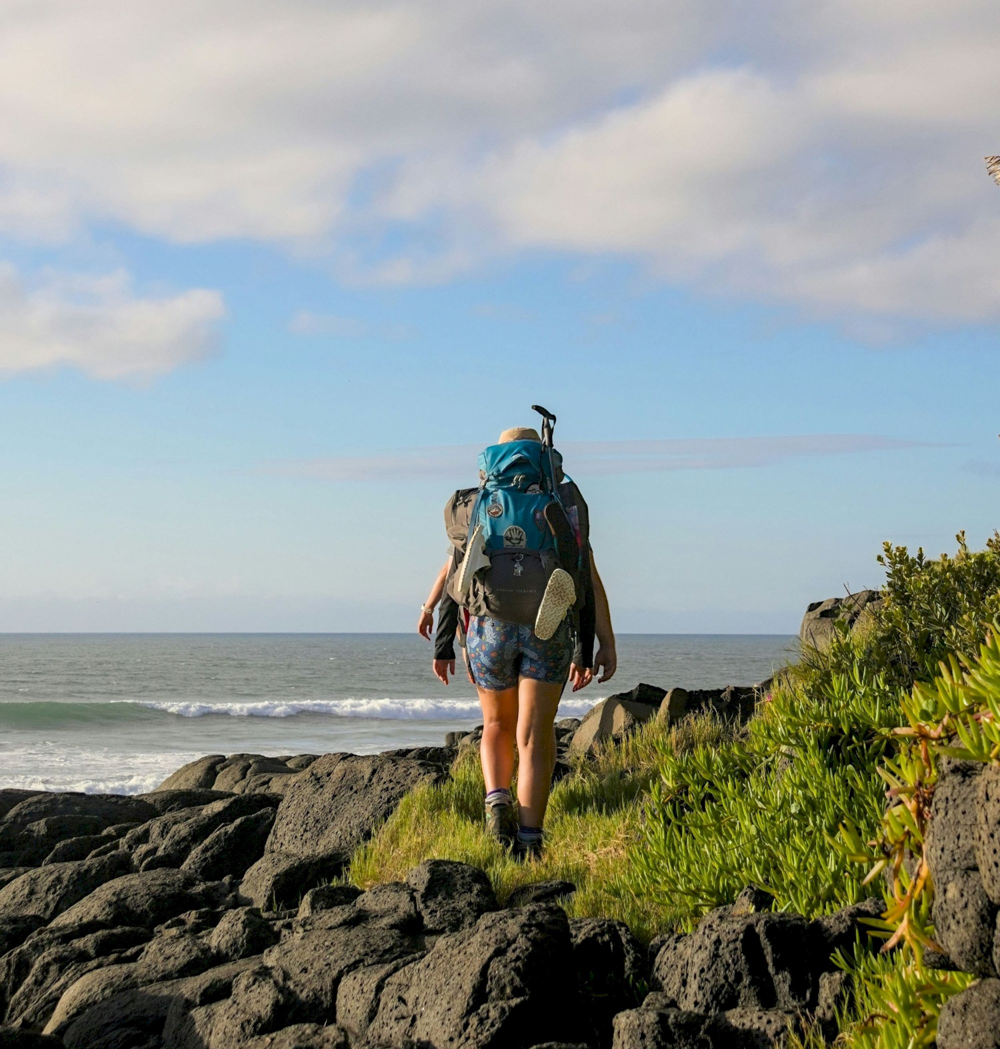 A hiker with a backpack walks along rocky coastline toward the sea, under a partly cloudy sky, wearing blue gear.