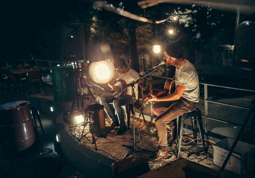 Two musicians perform a late-night acoustic set outdoors, playing guitars by soft stage lights on a small, intimate rooftop stage.