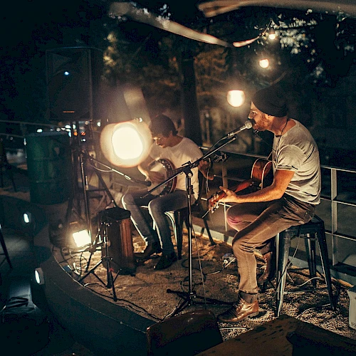 Two musicians perform a late-night acoustic set outdoors, playing guitars by soft stage lights on a small, intimate rooftop stage.