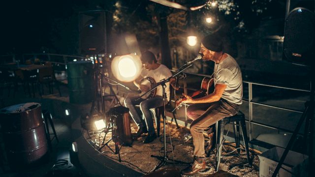 Two musicians perform a late-night acoustic set outdoors, playing guitars by soft stage lights on a small, intimate rooftop stage.