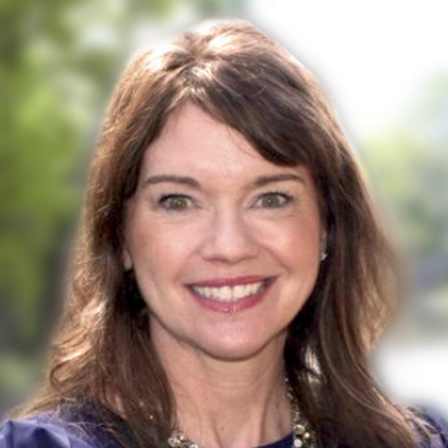 A smiling woman with shoulder-length brown hair, wearing a navy top and layered pearl necklace, outdoors.