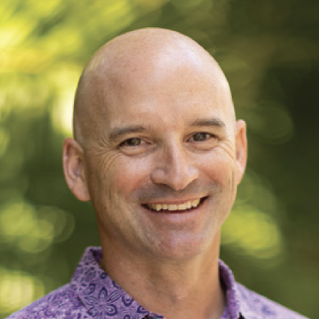 A smiling bald man wearing a purple patterned shirt outdoors, with a blurred green background.