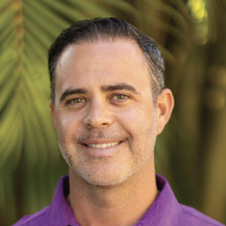 A smiling man in a purple polo shirt standing outdoors, with green palm leaves in the background.