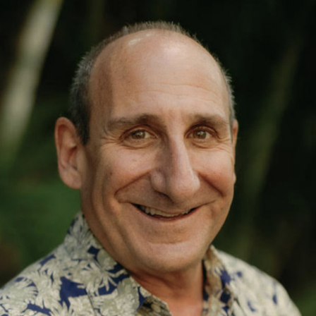 A smiling, balding man in a tropical-patterned shirt outdoors, with greenery in the background, looking friendly and warm.