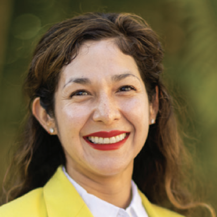 A smiling woman with long brown hair in a yellow jacket and white shirt, outdoors, with a warm, friendly expression.
