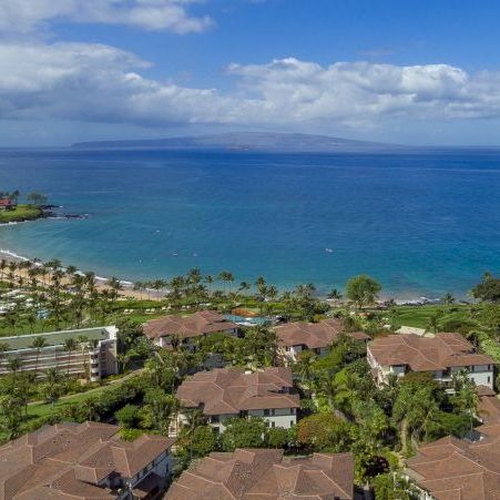 A tropical resort area along a curved coastline with brown-roofed villas, palm trees, and clear blue sea under a partly cloudy sky.