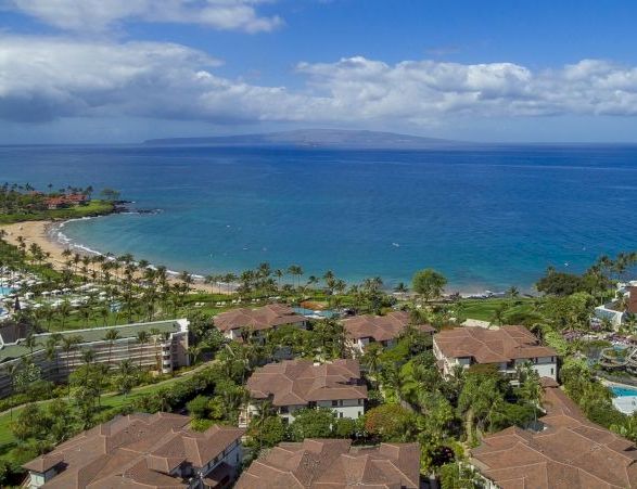 A tropical resort area along a curved coastline with brown-roofed villas, palm trees, and clear blue sea under a partly cloudy sky.