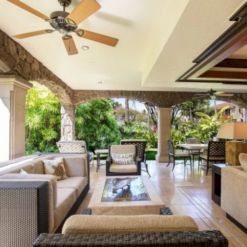 A bright, open outdoor living area with wicker and cushioned seating, a ceiling fan, stone columns, and a view of lush greenery and patio dining, all under a covered terrace.
