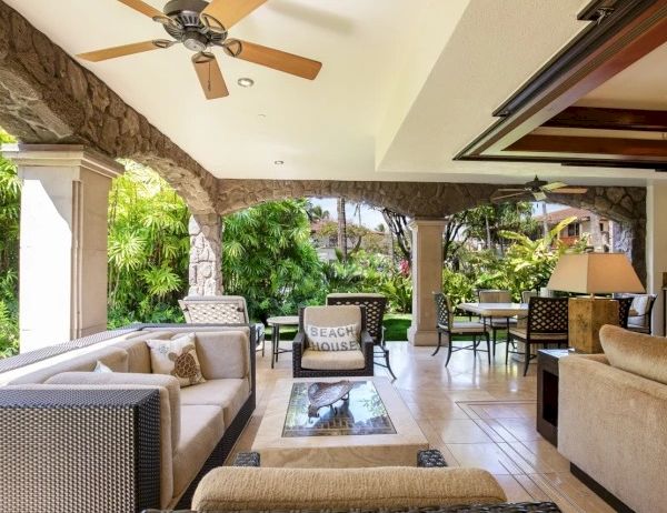 A bright, open outdoor living area with wicker and cushioned seating, a ceiling fan, stone columns, and a view of lush greenery and patio dining, all under a covered terrace.