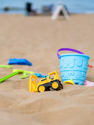 A sunny beach with sand toys scattered: a yellow toy truck, colorful shovels, a light blue bucket, and a purple-rimmed bucket lid by the shore.