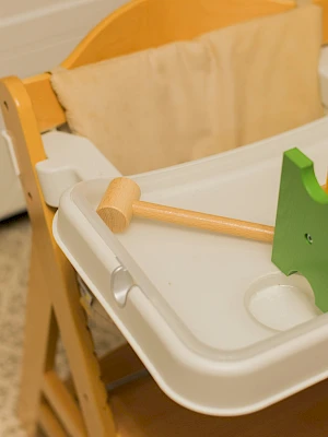 A wooden high chair with a white tray, a wooden mallet and a green peg block toy on the tray—kid-friendly kitchen play area.