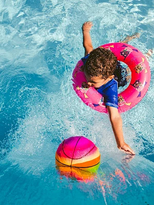 A child in a pink floatie reaches for a colorful ball in a clear blue swimming pool, with sunlight sparkling on the water.