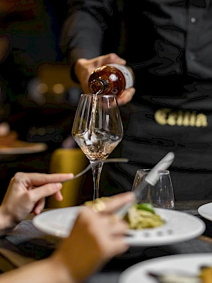 Someone pours red wine into a glass over a dining table with plates and hands nearby, in a stylish dining setting.