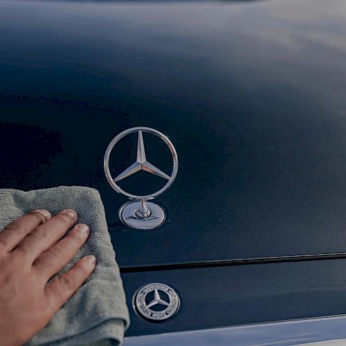 A hand wipes a dark Mercedes car hood, with three Mercedes logos visible on the hood and badge&mdash;clean, glossy, and branded.