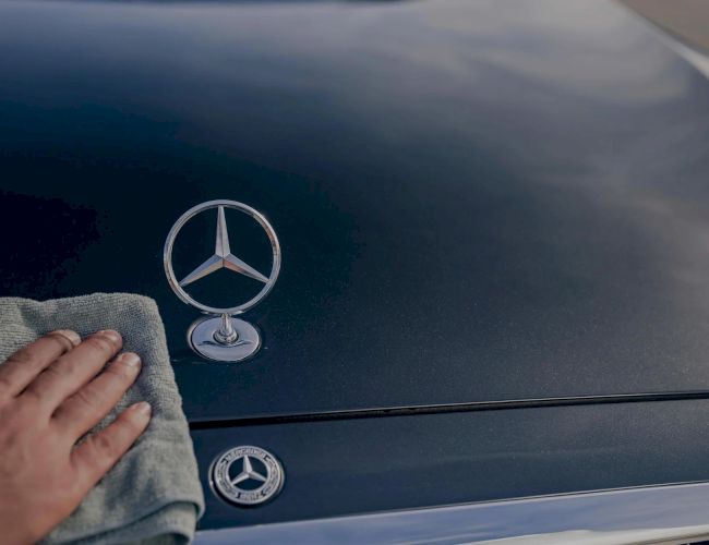 A hand wipes a dark Mercedes car hood, with three Mercedes logos visible on the hood and badge&mdash;clean, glossy, and branded.