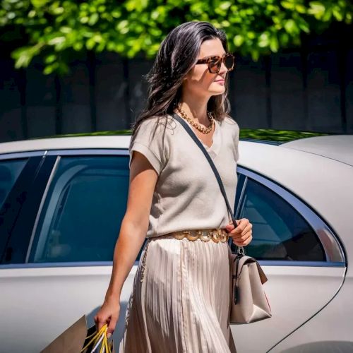 A woman wearing a beige top and pleated skirt with sunglasses walks beside a white car, holding a bag and shopping bags.