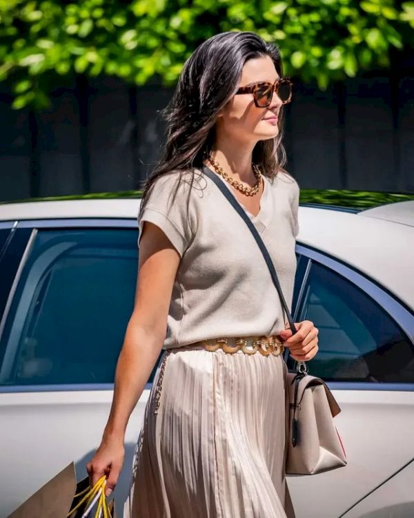 A woman wearing a beige top and pleated skirt with sunglasses walks beside a white car, holding a bag and shopping bags.