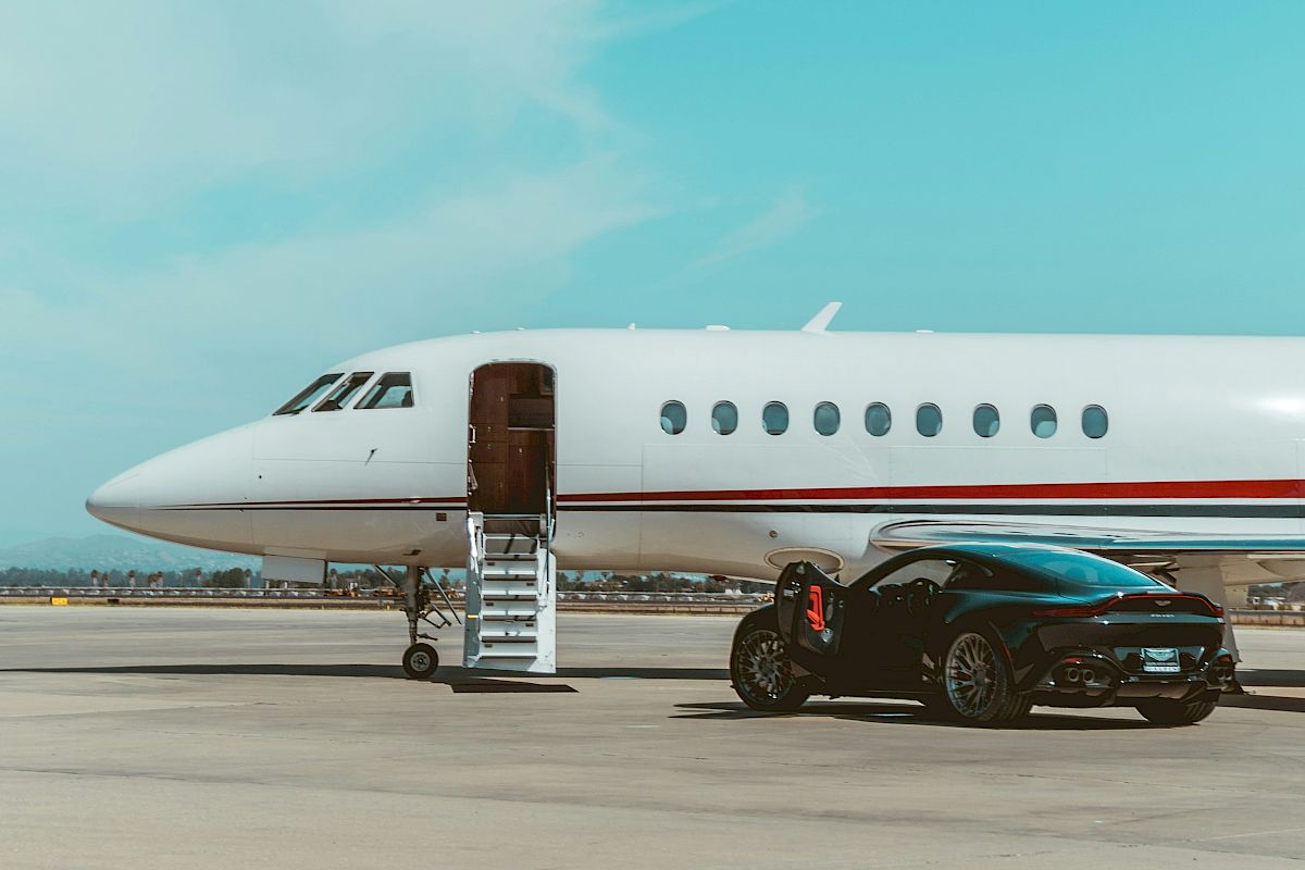 A sleek white private jet on a tarmac with an open door and a black car parked beside it under a bright, clear blue sky.