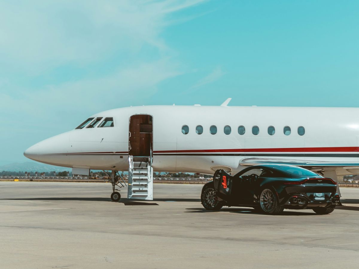 A sleek white private jet on a tarmac with an open door and a black car parked beside it under a bright, clear blue sky.