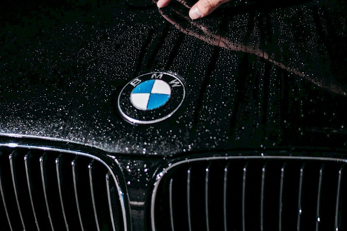 A hand wipes a wet, black BMW hood with a water spray shimmering on the surface.