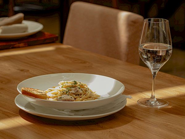 A plated dish with pasta sits on a wooden table, accompanied by a wine glass in a softly lit dining room, ready for a meal.